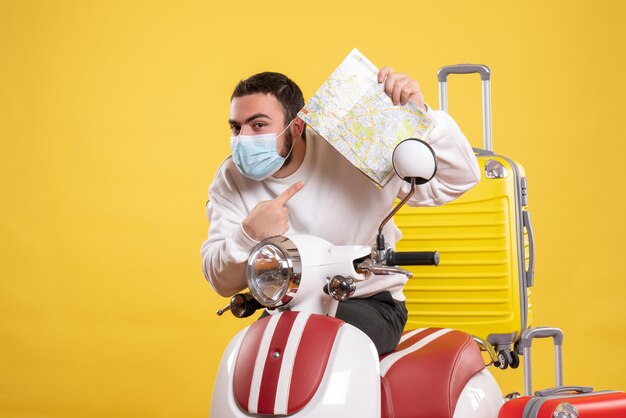 Vista superior del concepto de viaje con un chico joven con una máscara médica de pie cerca de la motocicleta con una maleta amarilla y sosteniendo un mapa sintiéndose curioso