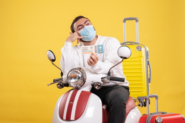 Vista superior del concepto de viaje con un chico joven con máscara médica sentado en una motocicleta con una maleta amarilla y sosteniendo un boleto haciendo un gesto de llamarme