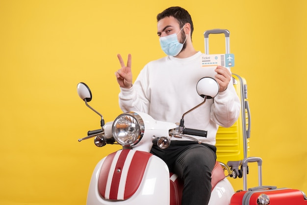 Vista superior del concepto de viaje con un chico joven con máscara médica sentado en una motocicleta con una maleta amarilla y sosteniendo un boleto haciendo un gesto de victoria