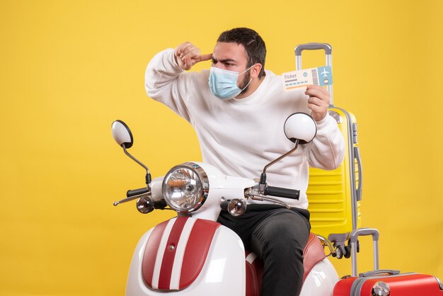 Vista superior del concepto de viaje con un joven pensador en máscara médica sentado en una motocicleta con una maleta amarilla y sosteniendo un boleto