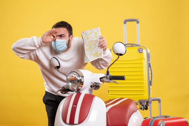 Vista superior del concepto de viaje con un tipo concentrado en una máscara médica de pie cerca de la motocicleta con una maleta amarilla y un boleto de retención