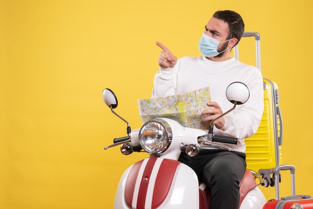 Vista superior del concepto de viaje con un tipo curioso con máscara médica sentado en una motocicleta con una maleta amarilla y sosteniendo un mapa