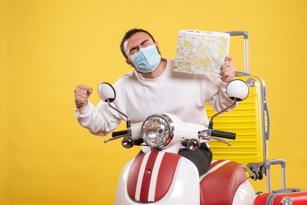 Vista superior del concepto de viaje con un tipo orgulloso y feliz en una máscara médica de pie cerca de la motocicleta con una maleta amarilla y sosteniendo el mapa
