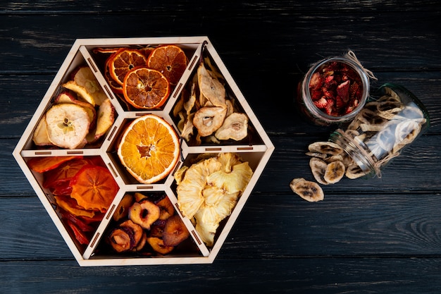 Foto gratuita vista superior de un conjunto de frutas secas en una caja de madera y plátano seco y chips de fresa en frascos de vidrio sobre fondo negro