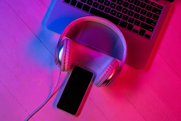 Vista superior del conjunto de gadgets en luz de neón púrpura y fondo rosa. Teclado portátil, auriculares y smartphone con pantalla negra. Copyspace para su publicidad. Tecnología, moderna, gadgets.