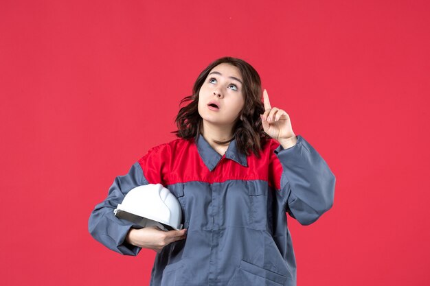 Vista superior del constructor femenino sorprendido en uniforme y sosteniendo el casco apuntando hacia arriba sobre fondo rojo aislado