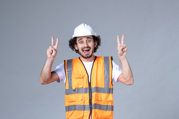 Vista superior del constructor masculino sonriente con chaleco de advertencia con casco de seguridad y haciendo gesto de victoria en la pared gris de la ola