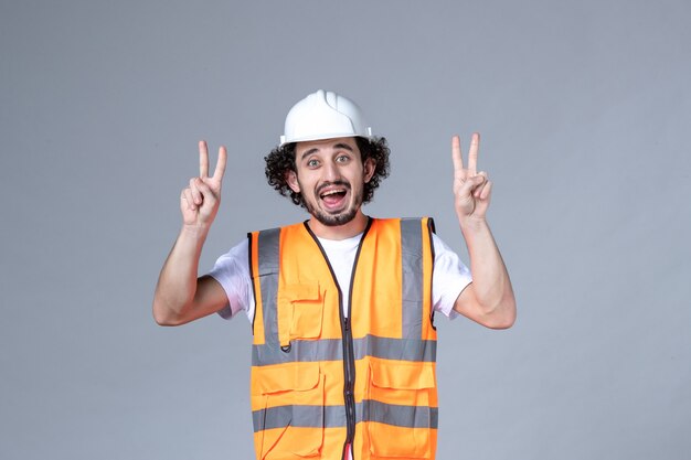 Vista superior del constructor masculino sonriente con chaleco de advertencia con casco de seguridad y haciendo gesto de victoria en la pared gris de la ola