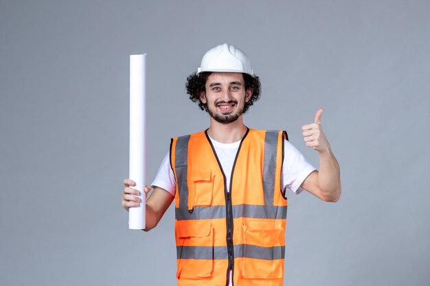 Vista superior del constructor masculino sonriente en chaleco de advertencia con casco de seguridad y sosteniendo un gesto en blanco haciendo bien en la pared de onda gris