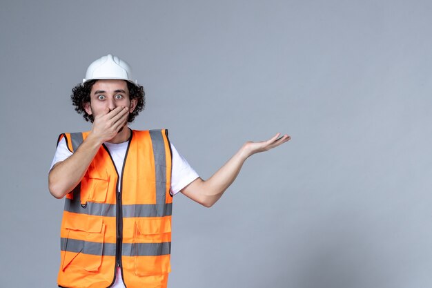 Vista superior del constructor masculino sorprendido en chaleco de advertencia con casco de seguridad y apuntando algo en el lado izquierdo sobre la pared gris de la ola