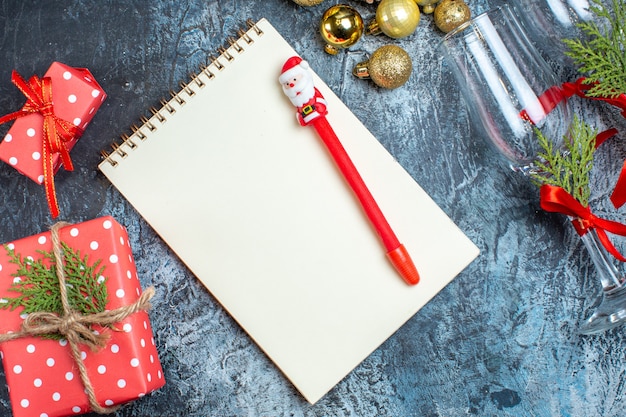 Vista superior de copas de cristal caídas con cinta roja y accesorios de decoración junto a cajas de regalo cuaderno con bolígrafo sobre fondo oscuro