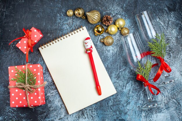 Vista superior de copas de cristal con cinta roja y accesorios de decoración junto a cajas de regalo cuaderno con bolígrafo sobre fondo oscuro