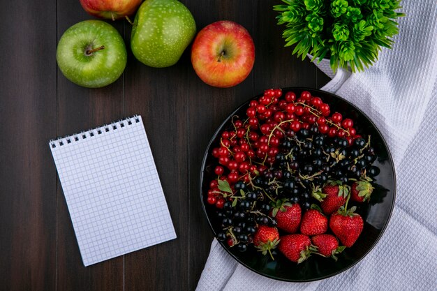 Vista superior copia espacio grosellas rojas y negras con fresas en un plato con manzanas y un cuaderno sobre un fondo de madera