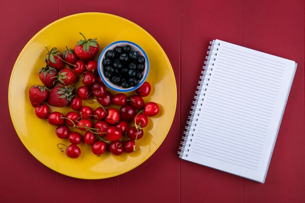 Vista superior copia espacio portátil con arándanos, fresas y cerezas en un plato sobre un fondo rojo.