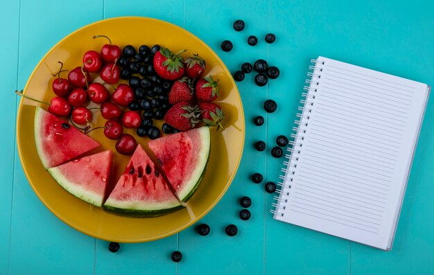 Vista superior copia espacio portátil con rodajas de sandía fresas cerezas y arándanos en un plato amarillo sobre un fondo azul claro