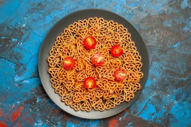 Vista superior corazones de pasta italiana cortan tomates cherry en placa ovalada negra sobre mesa azul