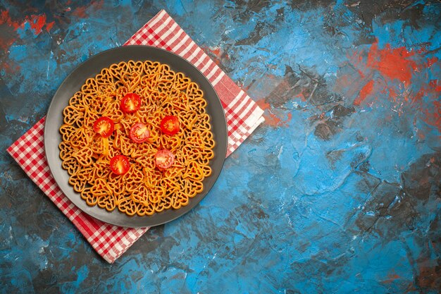 Vista superior corazones de pasta italiana cortan tomates cherry en un plato sobre una toalla de cocina a cuadros roja blanca sobre una mesa azul con espacio libre