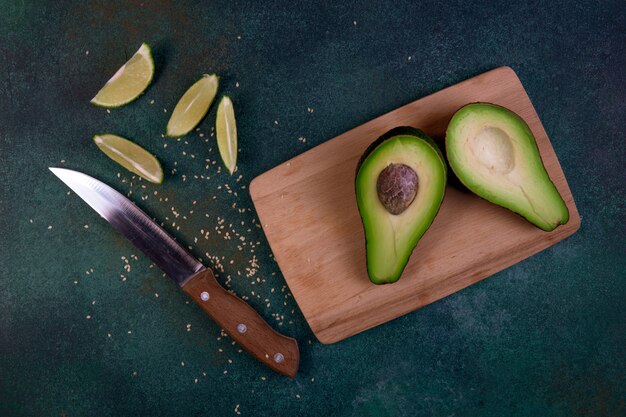 Vista superior cortada por la mitad aguacate en una pizarra con limón y cuchillo sobre un fondo verde oscuro