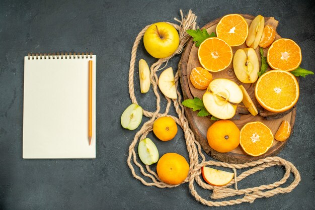 Vista superior cortar manzanas y naranjas en tablero de madera cóctel un cuaderno y lápiz sobre fondo oscuro