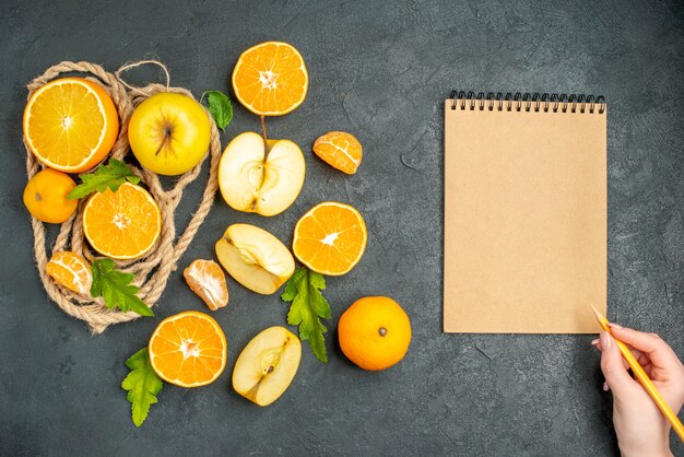 Vista superior cortar naranjas y manzanas con un lápiz de bloc de notas en mano femenina sobre fondo oscuro