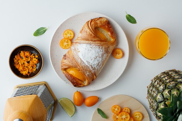 Vista superior de croissant con rodajas de kumquat en plato y jugo de naranja de ralladura de naranja de piña con rallador sobre fondo blanco