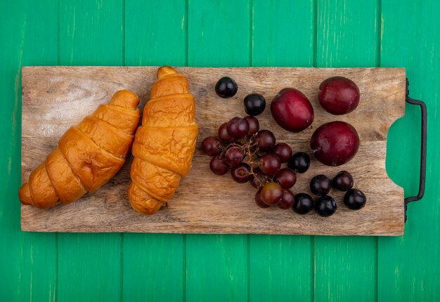 Vista superior de croissants y bayas de endrinas pluots de uva en tabla de cortar sobre fondo verde