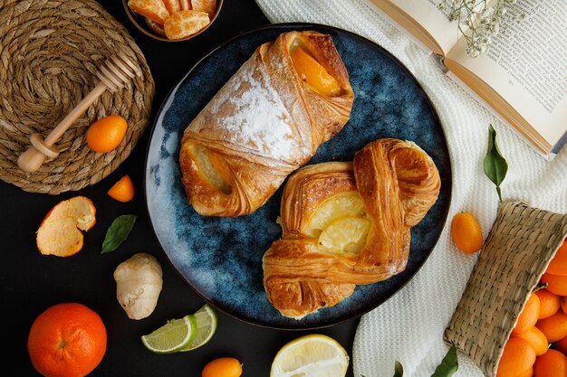 Vista superior de croissants en plato con rodajas de mandarina con concha y libro abierto con cesta de kumquat y florecita en libro con trivet de rodajas de jengibre y cucharón de miel sobre tela