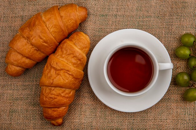 Vista superior de croissants y taza de té con uva sobre fondo de tela de saco