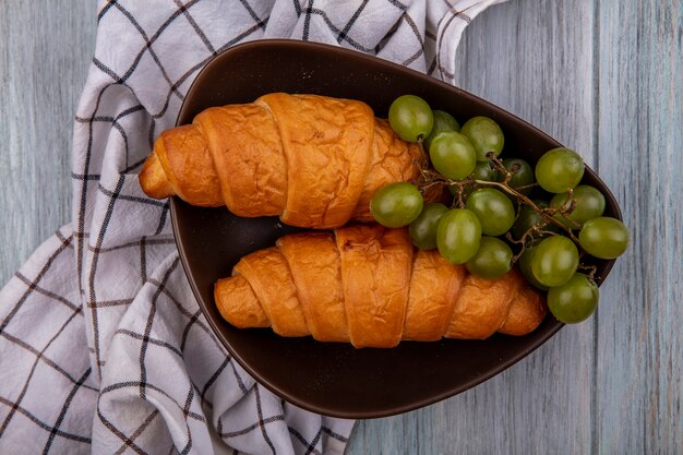 Vista superior de croissants y uva en un tazón sobre tela escocesa sobre fondo de madera