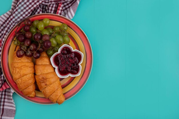 Vista superior de croissants con uvas y mermelada de frambuesa en placa sobre tela escocesa sobre fondo azul con espacio de copia