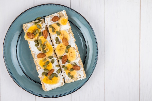 Vista superior de crujientes galletas con queso crema, rodajas de plátano, almendras y semillas de calabaza en un plato sobre madera blanca con espacio de copia