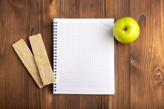 Vista superior del cuaderno azul abierto con galletas y manzana verde en el escritorio marrón