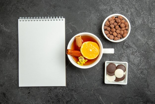 Vista superior del cuaderno blanco de hizelnuts de chocolate junto a los cuencos de chocolate y hizelnuts y una taza de té con canela y limón en la superficie oscura