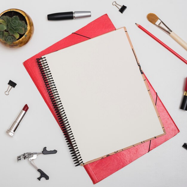 Vista superior de cuaderno en blanco con productos cosméticos; clips de papel; lapiz sobre escritorio blanco