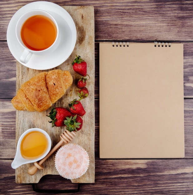 Vista superior de un cuaderno de bocetos y fresas maduras frescas croissant miel y una taza de té en una tabla para cortar madera rústica