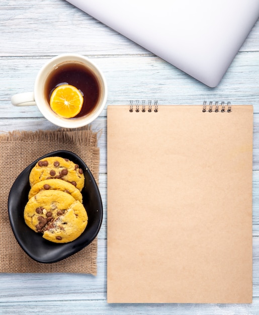 Vista superior del cuaderno de bocetos y galletas de avena con chispas de chocolate y una taza de té en rústico