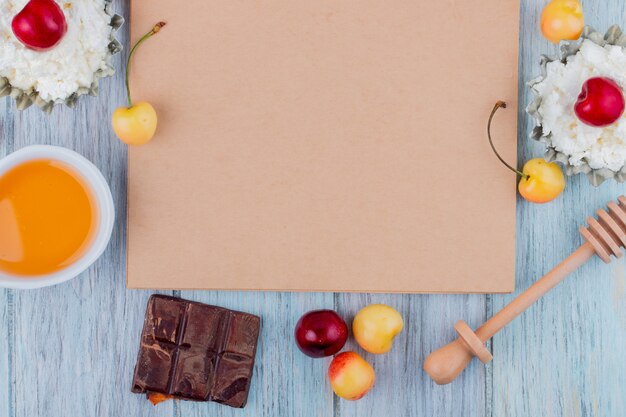 Vista superior del cuaderno de bocetos y queso cottage con miel de chocolate negro y cerezas rojas y amarillas maduras frescas dispuestas en gris
