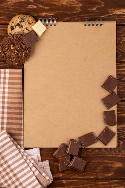 Vista superior del cuaderno de bocetos con trozos de chocolate negro y galletas de avena sobre fondo de madera