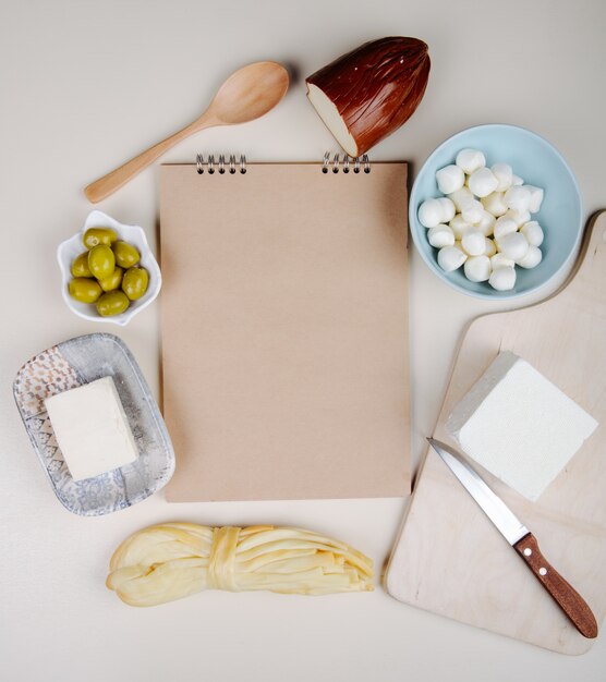 Vista superior de un cuaderno de bocetos y varios tipos de queso mini queso mozzarella en un tazón azul, queso feta, ahumado y queso con aceitunas en vinagre en mesa blanca