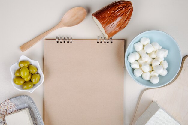 Vista superior de un cuaderno de bocetos y varios tipos de queso mini queso mozzarella en un tazón azul, queso feta, ahumado y queso con aceitunas en vinagre en mesa blanca