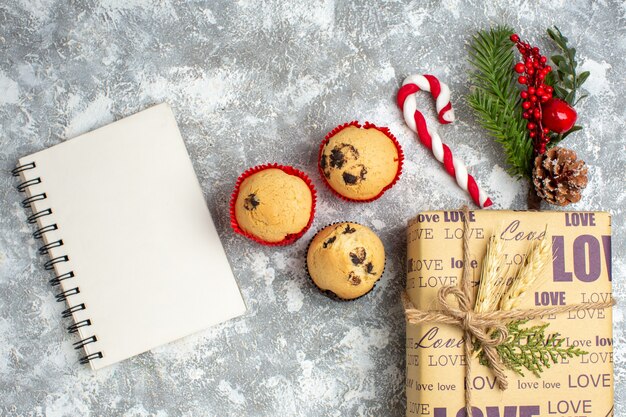 Vista superior del cuaderno cerrado y pequeños cupcakes hermoso regalo de Navidad con inscripción de amor y accesorios de decoración de ramas de abeto, cono de coníferas en el lado izquierdo sobre la superficie del hielo