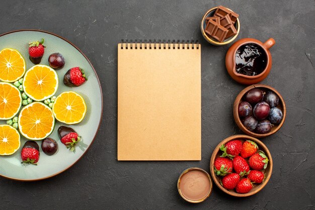 Vista superior del cuaderno de crema de salsa de chocolate entre fresas de chocolate en tazones y plato de pastel con salsa de chocolate sobre fondo negro