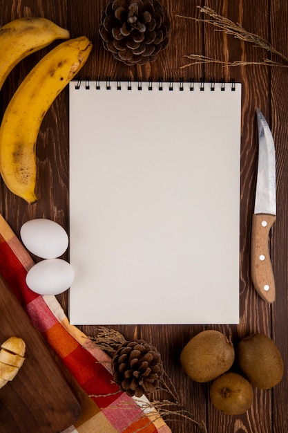 Vista superior de un cuaderno de dibujo con kiwi y plátano y dos huevos con cuchillo de cocina en madera