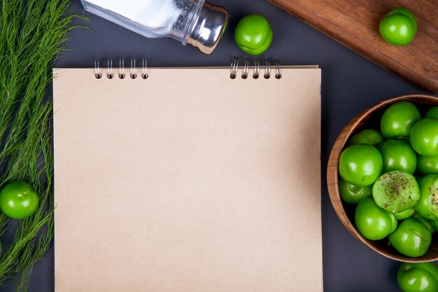 Vista superior de un cuaderno de dibujo, salero, hinojo y ciruelas verdes agrias en un tazón de madera sobre una mesa negra