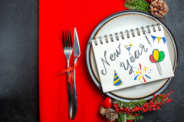 Vista superior del cuaderno con escritura de año nuevo y dibujos en un plato con accesorios de decoración, ramas de abeto y cubiertos en una servilleta roja