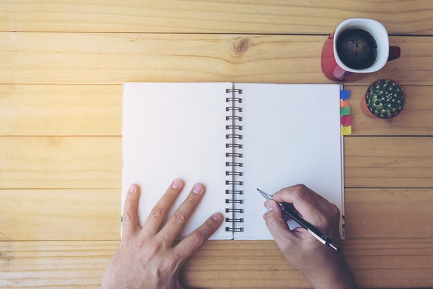 Vista superior del cuaderno de escritura de hombre en mesa de madera con taza de café y olla pequeña de cactus