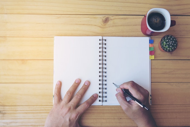 Vista superior del cuaderno de escritura de hombre en mesa de madera con taza de café y olla pequeña de cactus