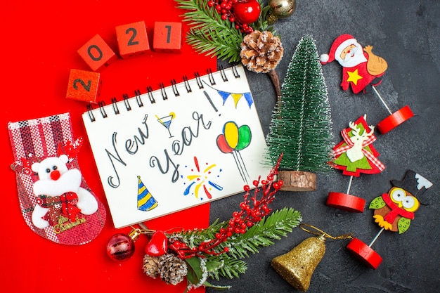 Vista superior del cuaderno espiral con escritura de año nuevo y dibujos, accesorios de decoración, ramas de abeto, números de calcetines navideños en una servilleta roja y árbol de Navidad sobre fondo oscuro