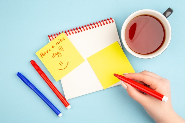 Vista superior del cuaderno espiral taza de té en la mano de la mujer que tenga un buen día escrito en marcadores de papel de nota en azul