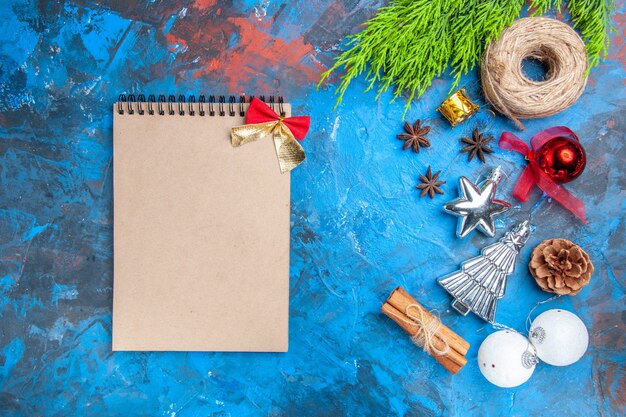 Vista superior de un cuaderno con pequeño lazo hilo de paja juguetes de árbol de navidad semillas de anís palitos de canela sobre fondo azul-rojo